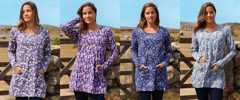 Four women standing outdoors in front of a wooden fence and stone wall, each wearing a long-sleeve tunic dress with a different floral print. The dresses feature purple, blue, and light blue floral patterns, and the background shows a rural landscape under a blue sky.