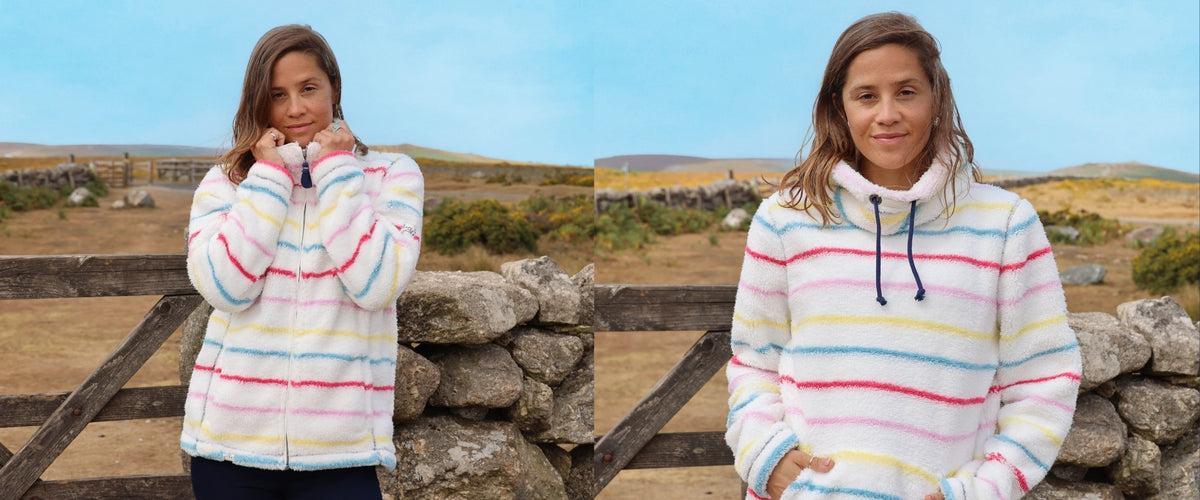 A side-by-side comparison of a woman wearing a pastel striped fleece zip-up jacket on the left and a matching high-neck fleece hoodie on the right, set against an outdoor rural background with a stone wall and blue sky.