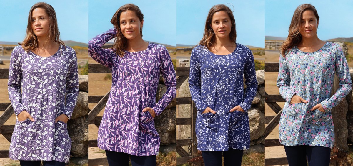 Four women standing outdoors in front of a stone wall and blue sky, each wearing a long-sleeve floral tunic top in a different colorway: purple/white, lavender/purple, blue/white, and multicolor pastel.