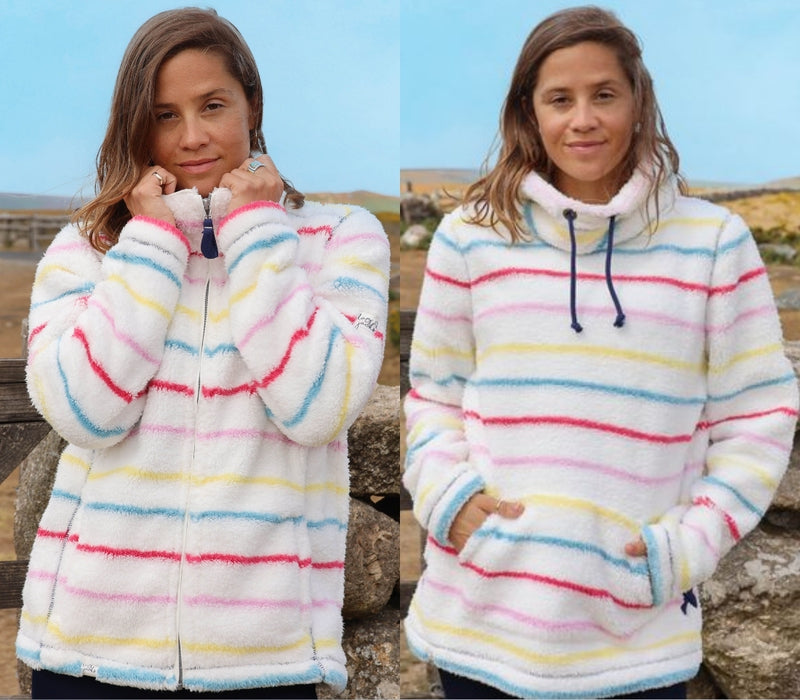 A woman modeling a fuzzy white fleece zip-up hoodie with pastel pink, blue, yellow, and green horizontal stripes, shown in a side-by-side front view. The background is an outdoor stone wall under a blue sky.