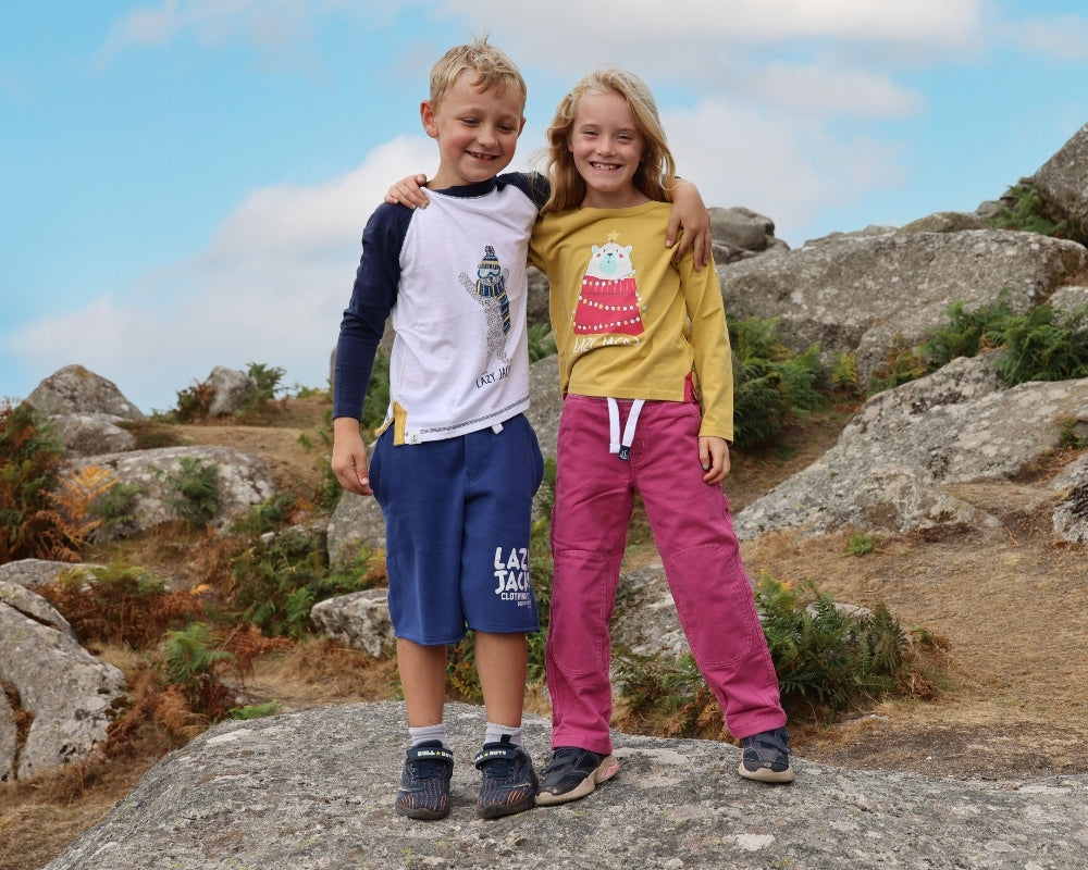Two children standing outdoors on a large rock in a rocky, grassy landscape under a blue sky. The boy wears a white and navy long-sleeve shirt with a graphic, navy shorts, and dark sneakers.
