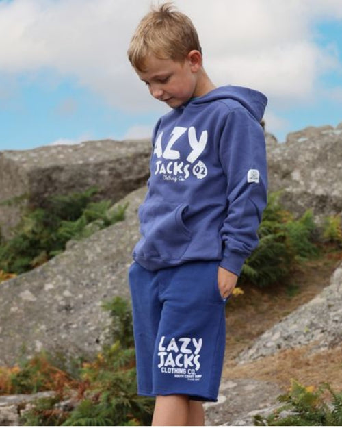 A young boy wearing a blue Lazy Jacks hoodie and matching shorts with white text, standing outdoors on a rocky hillside under a cloudy sky.